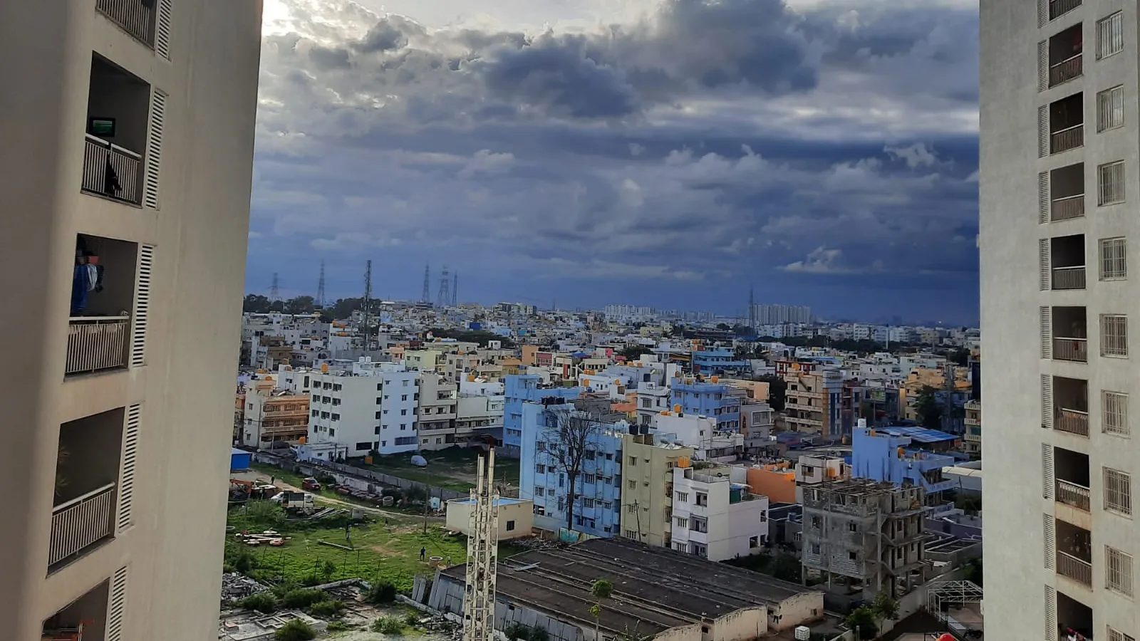 A dense Bengaluru residential neighbourhood of low-rise homes and apartment blocks, seen between two high-rise towers under monsoon clouds