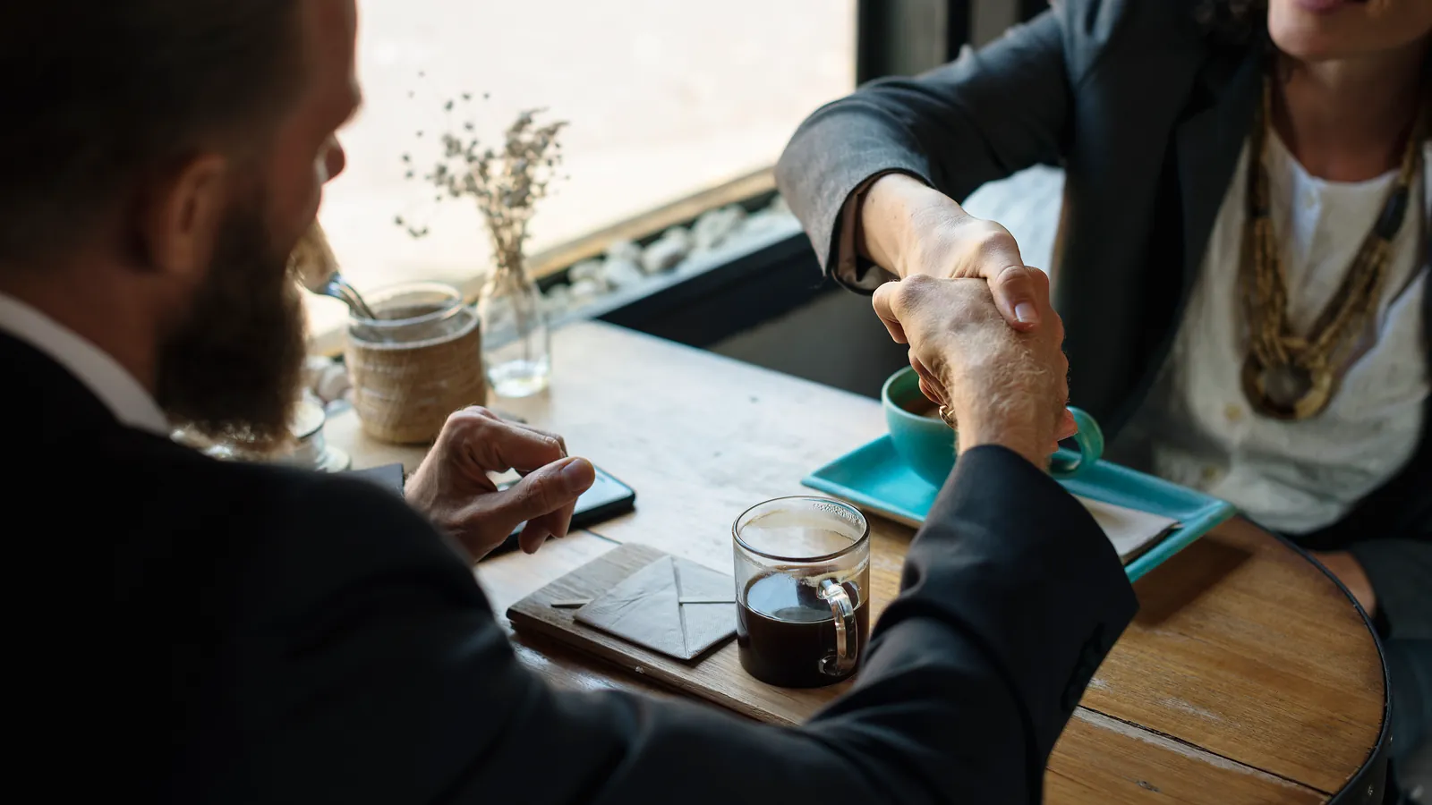 Two people shaking hands across a table with papers and coffee cups after reviewing an agreement
